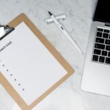 Caiet cu o listă de contacte de presă lângă un laptop, pe biroul unui specialist în comunicare